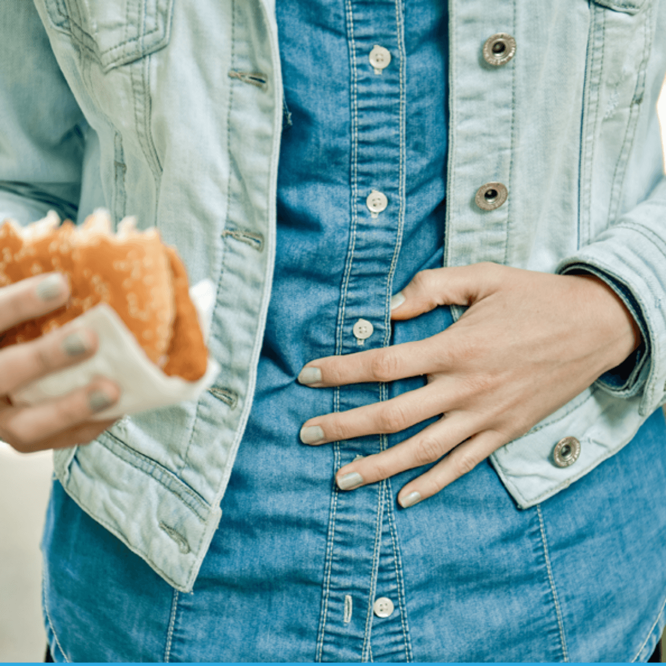 A close-up of a person standing, grabbing their stomach uncomfortably with one hand and holding a fried chicken sandwich in their other hand.