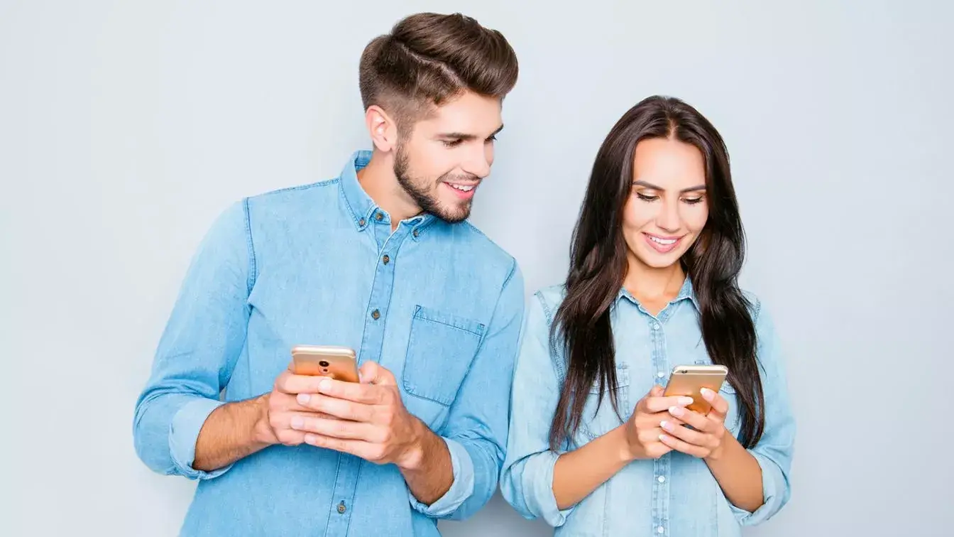 A man and a woman in denim shirts standing against a light blue wall, both looking at the woman's phone.