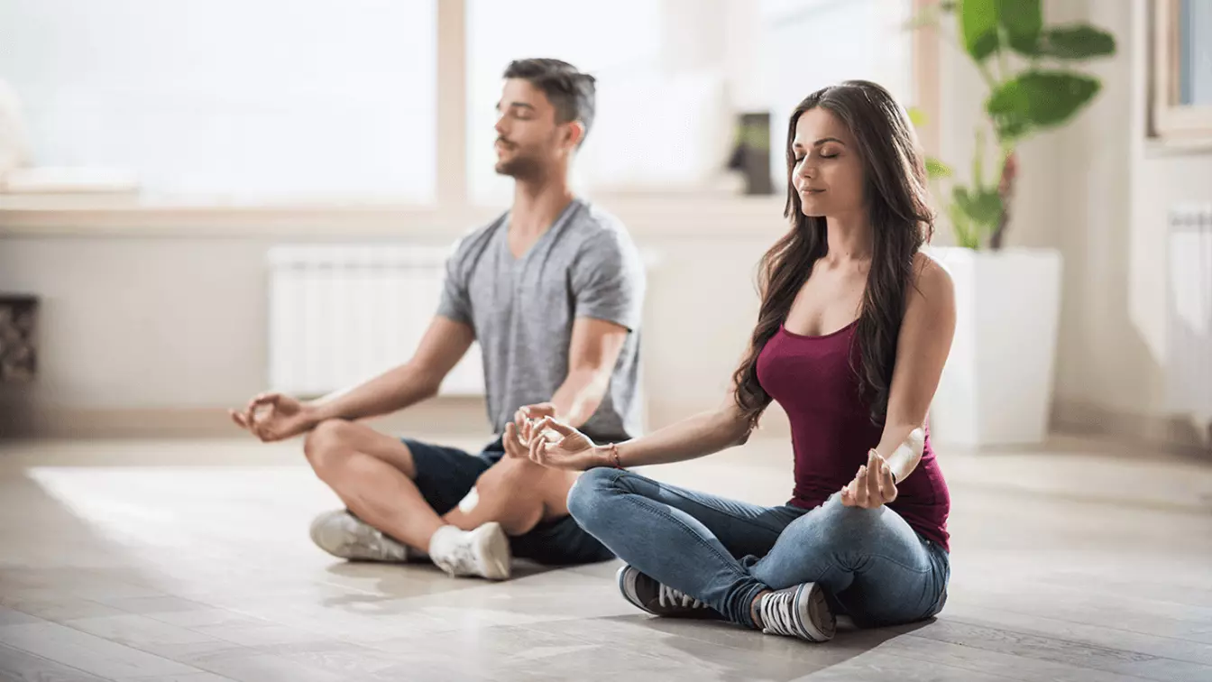 A woman and a man meditating in a room, sitting on the floor with their legs crossed and hands resting on their knees.